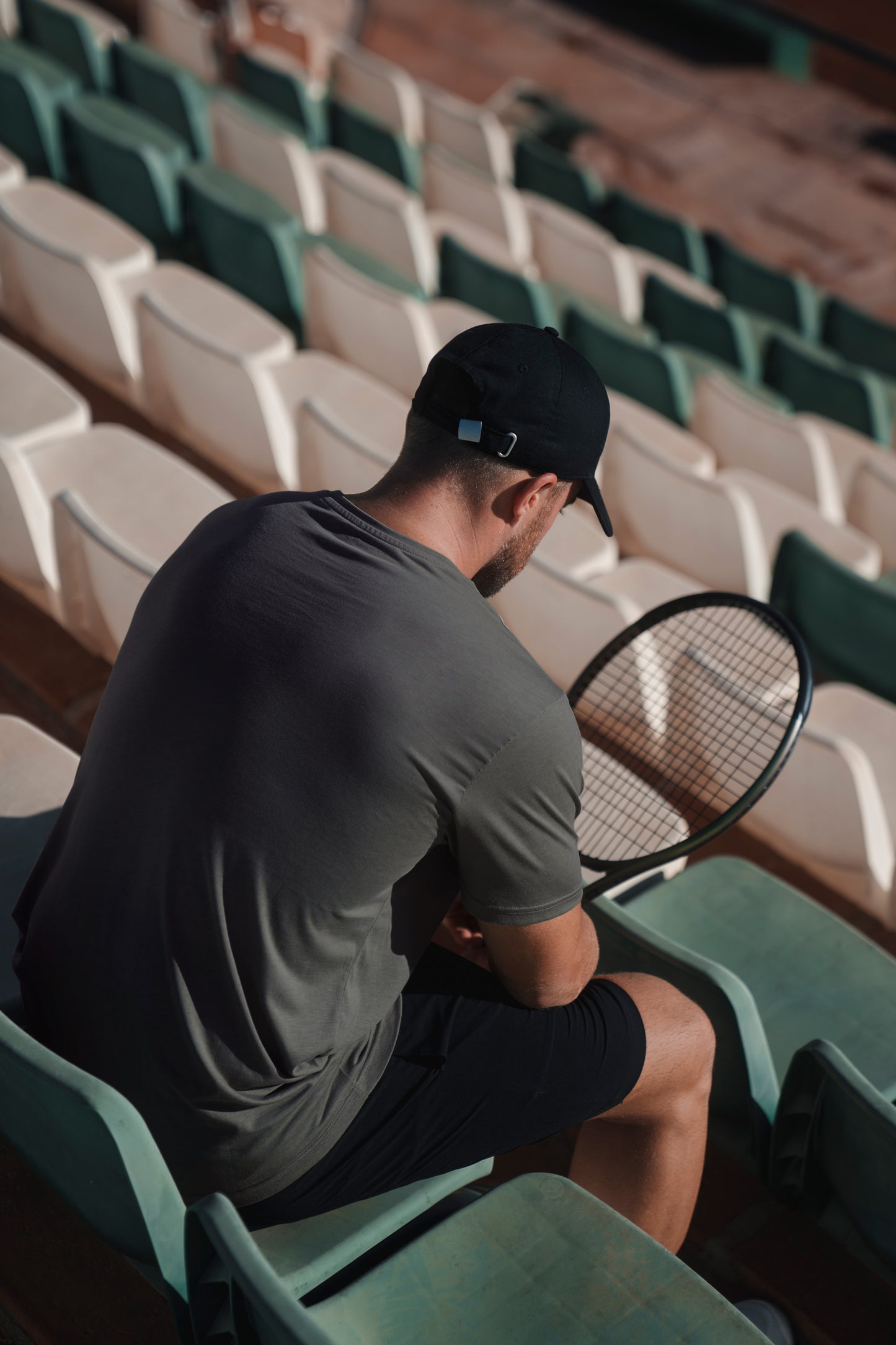 Man sitting on a tennis court with a racket, wearing a cap and asphalt lyocell t-shirt
