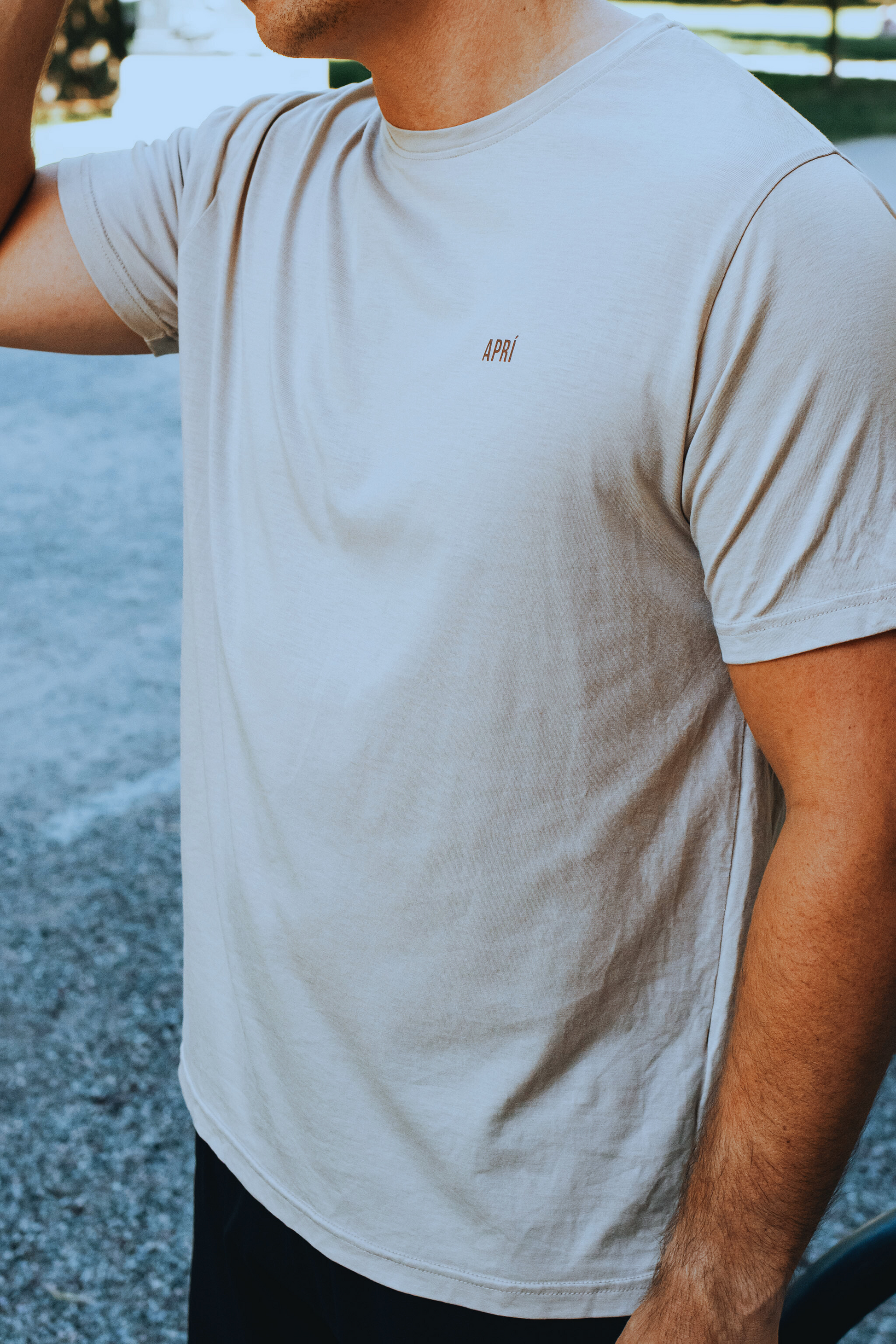 Person wearing a light gray t-shirt with a brand logo, standing outdoors.
