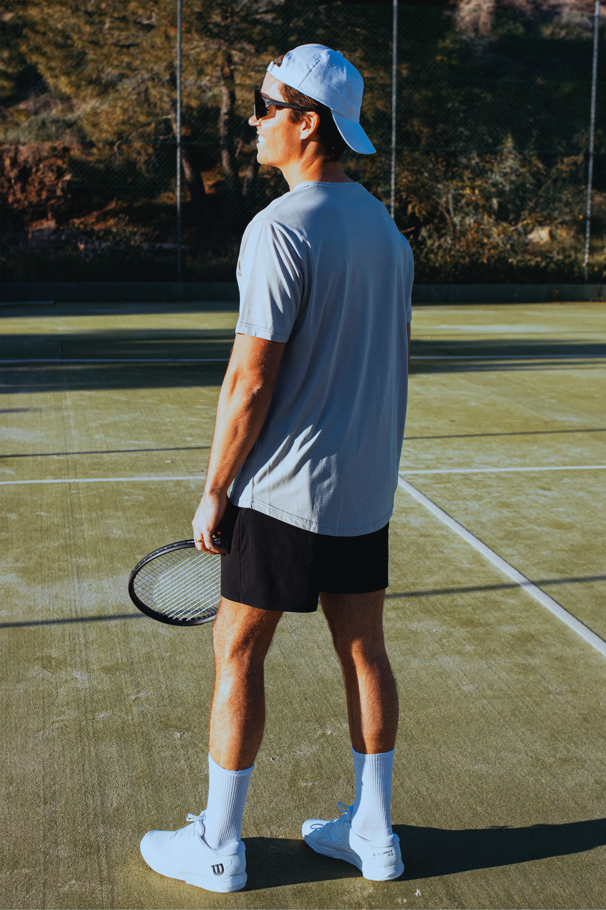 Person standing on a tennis court holding a racket, wearing a light blue shirt, black shorts, and white shoes.